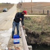 A scientist lowers a water sampling device off of a bridge into a rural stream