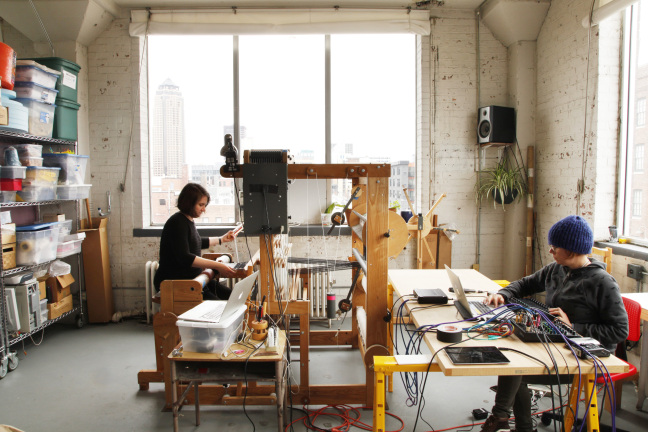 Olivia Valentine sits at a large wooden loom and weaves fabric while Paula Matthusen sits at right in front of a computer composing music.