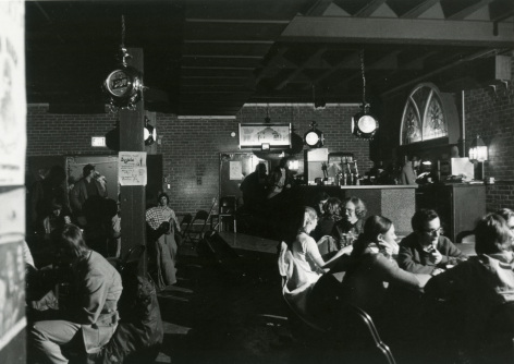 Black and white historical photo of patrons sitting in front of the bar of the Maintenance Shop