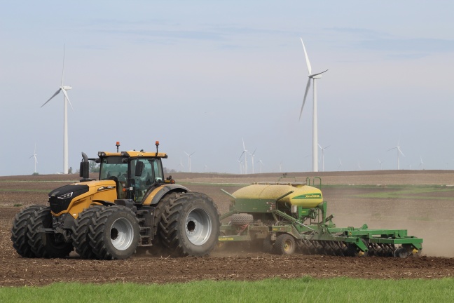 A yellow tractor hauls a green planter across a bare soil field, with wind turbines and an overcast sky in the background.