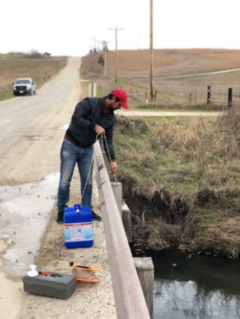 A scientist lowers a water sampling device off of a bridge into a rural stream