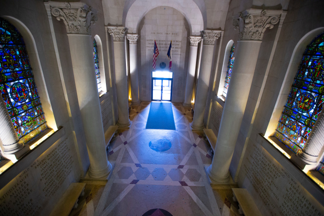 A photo of the Gold Star Hall at Iowa State University's Memorial Union. Gray stone pillars line the hall, and an American and Iowa flag stand against the far wall. The walls on the left and right are inscribed with the nemes of fallen veterans who are for