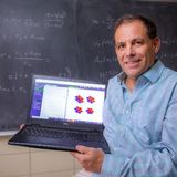 A photo showing theoretical physicist Alex Travesset in front of a blackboard full of equations and holding a laptop showing scientific figures.