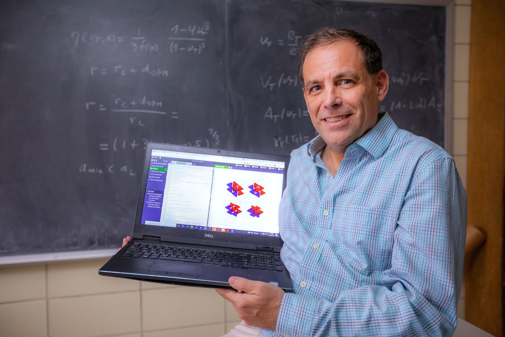 A photo showing theoretical physicist Alex Travesset in front of a blackboard full of equations and holding a laptop showing scientific figures.