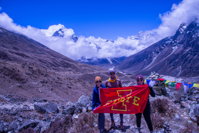 An Iowa State University professor and two students stand on a pass high in the Himalayas with mountain peaks in the background. They hold an Iowa State University flag.