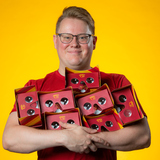 An ISU student smiles and holds seven virtual reality headsets in his arms. The headsets are cardboard and colored cardinal and gold.