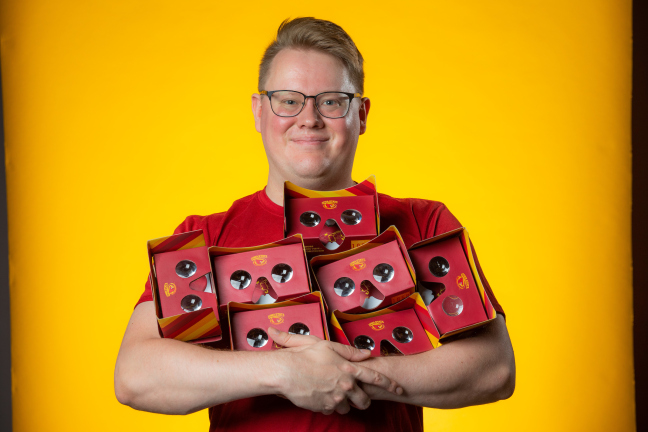 An ISU student smiles and holds seven virtual reality headsets in his arms. The headsets are cardboard and colored cardinal and gold.