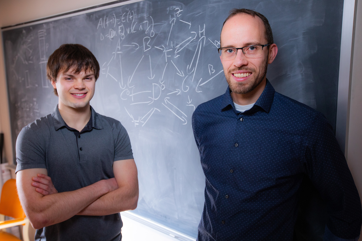 Iowa State senior Addison Schmidt (left) and associate professor of mathematics Claus Kadelka stand in front of a chalk board with depictions of a gene regulatory network in Carver Hall. Photo by Christopher Gannon/Iowa State University.