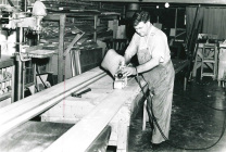 A black and white photo of a carpenter working on a wooden plank inside a cluttered workshop