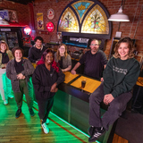 A handful of ISU students who work at the Maintenance Shop gather at the venue's bar and smile at the camera. The venue's iconic stained-glass window glows in the background.