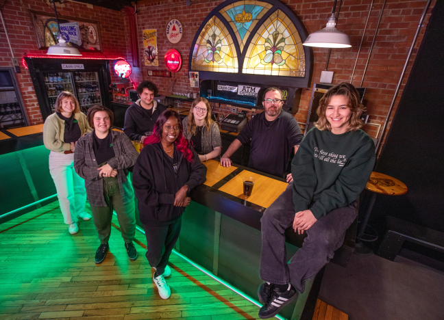 A handful of ISU students who work at the Maintenance Shop gather at the venue's bar and smile at the camera. The venue's iconic stained-glass window glows in the background.