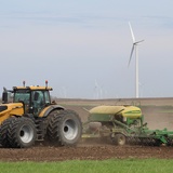 A yellow tractor hauls a green planter across a bare soil field, with wind turbines and an overcast sky in the background.