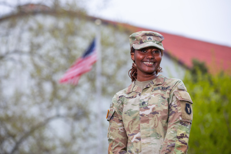 An ISU student smiles and wears National Guard fatigues. An American flag waves in the background.