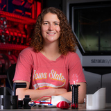 An ISU student smiles at the camera in a laboratory. On a table in front of her are several 3D-printed tools.