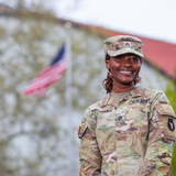 An ISU student wears National Guard fatigues and smiles at the camera. An American flag waves in the background.