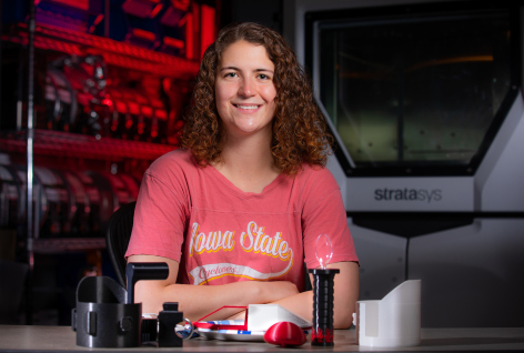An ISU student smiles at the camera in a lab. On a table in front of her are several 3D printed tools.